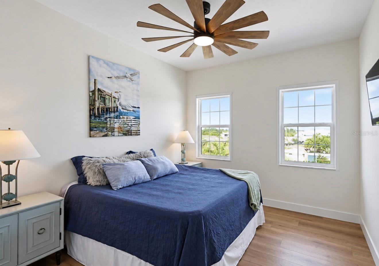 Furnished bedroom with navy bedding, coastal bird artwork, and a wood-blade ceiling fan.