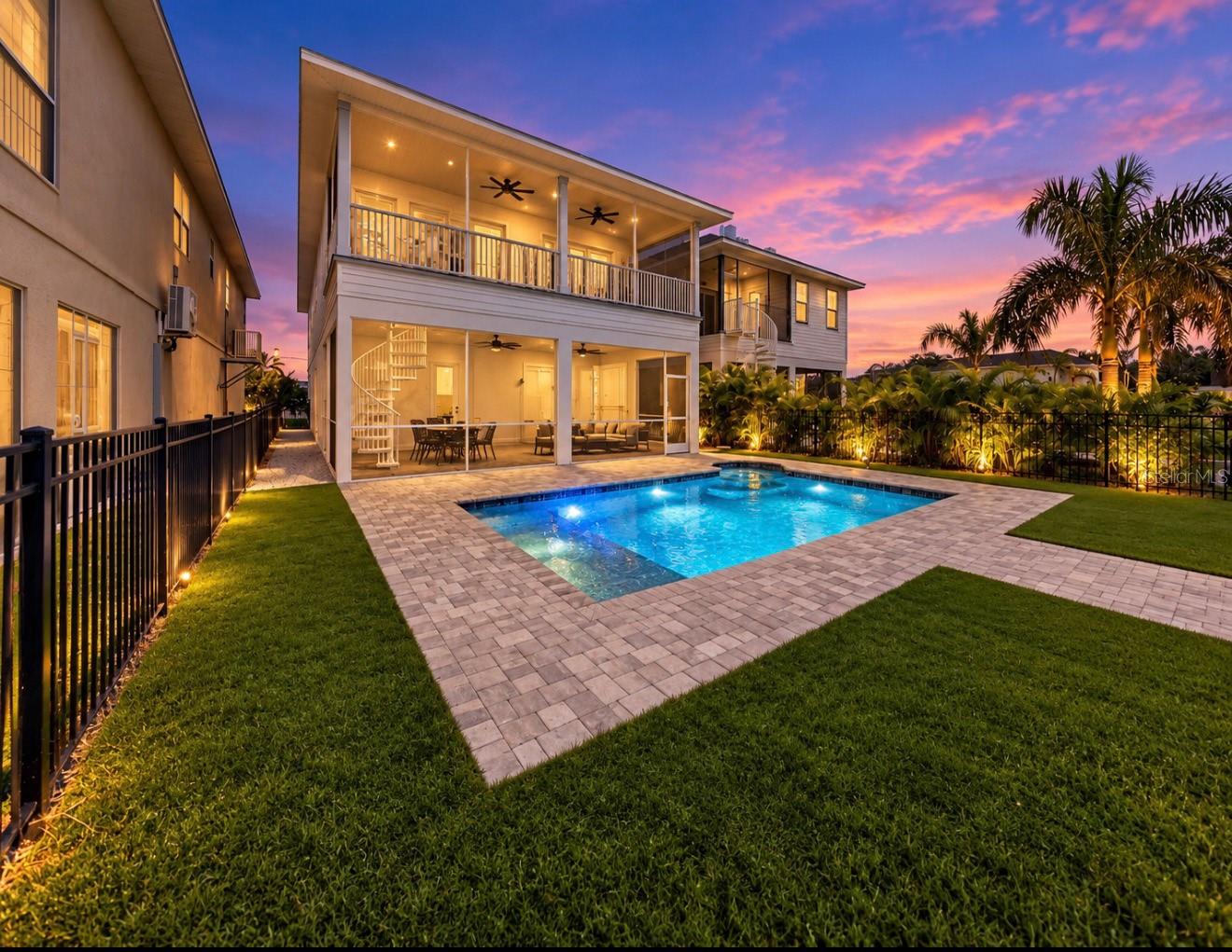 Two-story home with a lit pool, covered patio, and balcony beneath a pink and purple dusk sky.