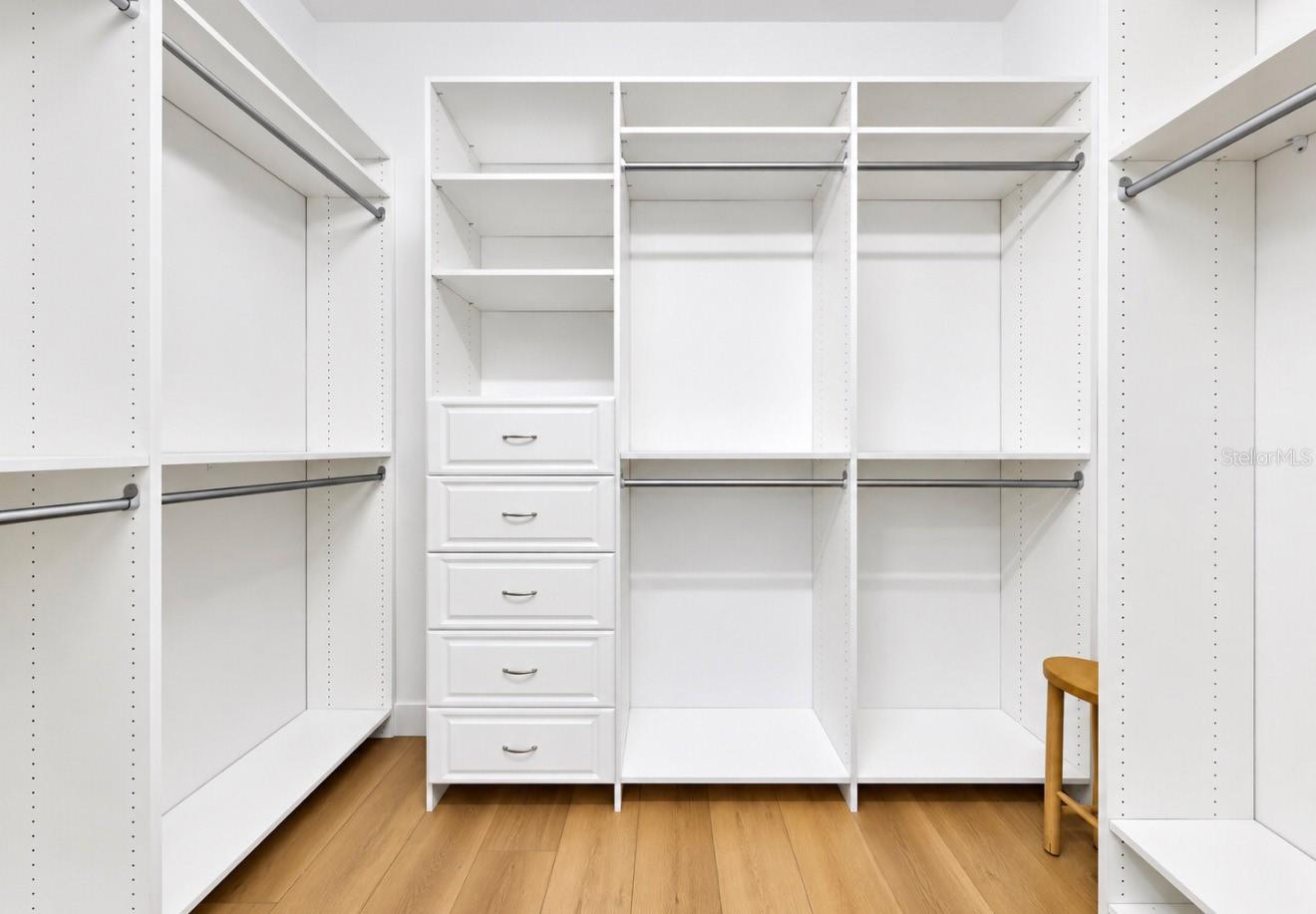 Walk-in closet with white built-in shelving, hanging rods, central drawers, and light wood flooring.