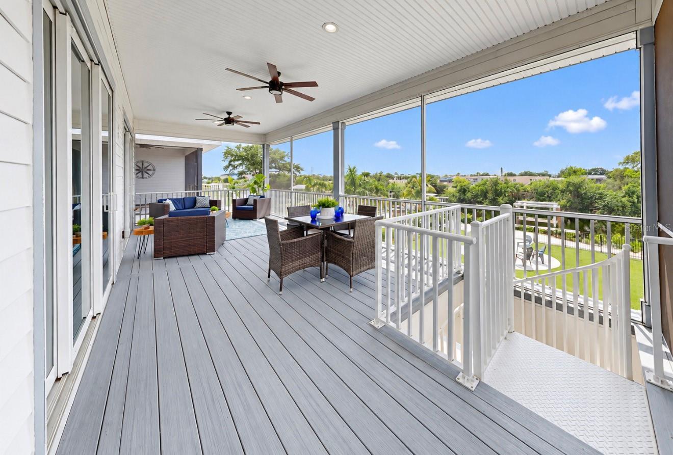 Covered balcony with lounge and dining furniture, gray decking, ceiling fans, and views of trees and homes.
