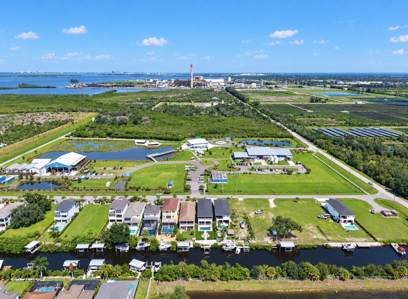Aerial view of waterfront homes along a canal, with wetlands and an industrial facility beyond.