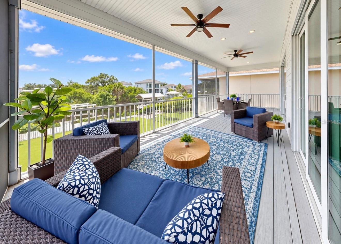 Screened lanai with blue-cushioned wicker seating, patterned rug, and views of trees and neighboring homes.