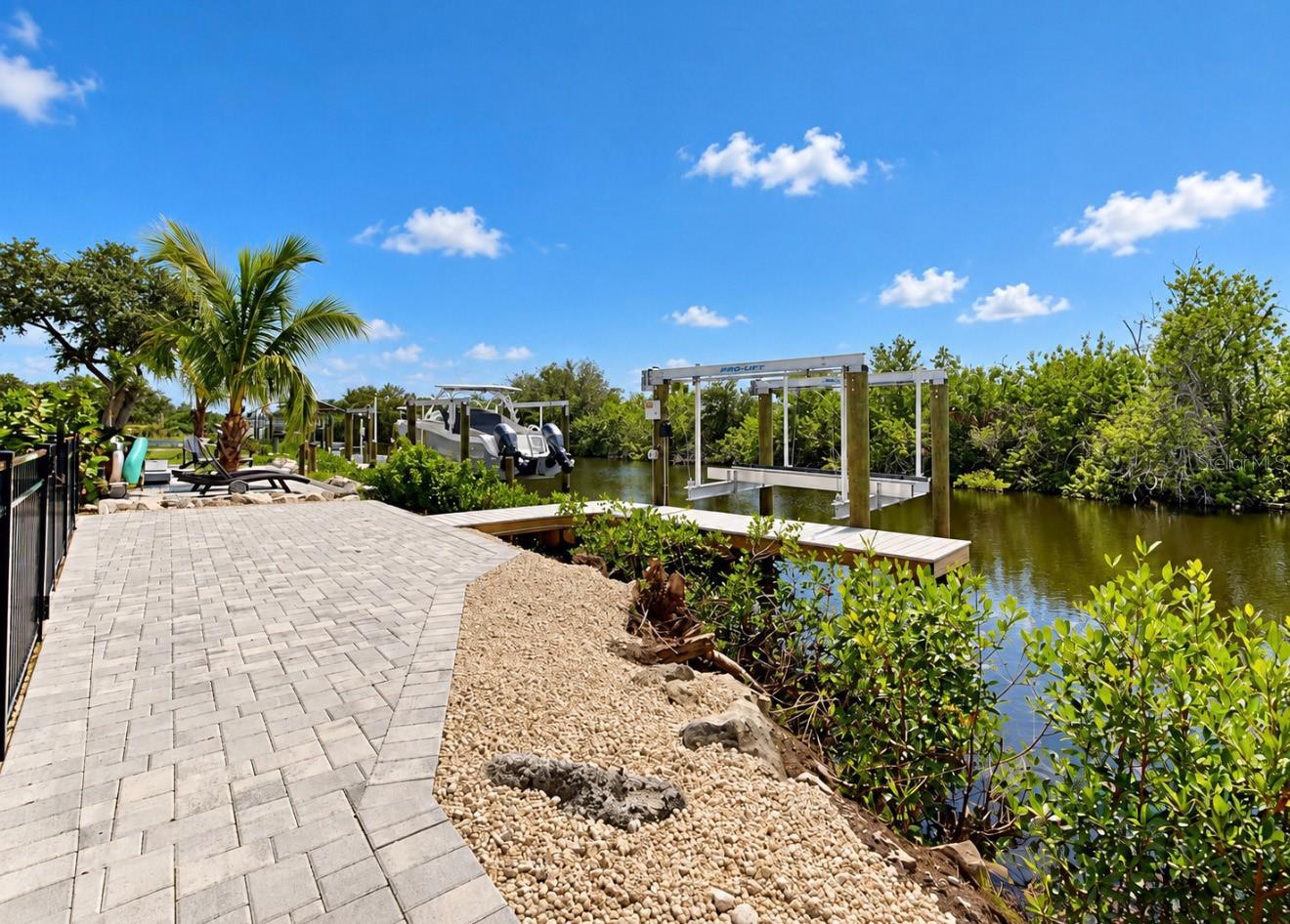 Canal-side dock with a boat lift beside a paver walkway and tropical landscaping.