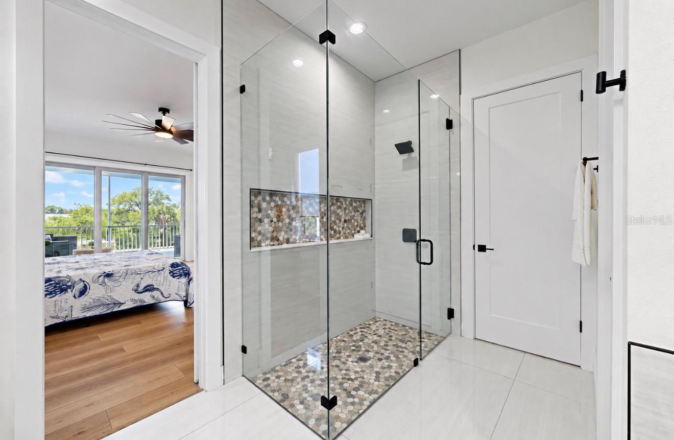Modern glass walk-in shower with pebble-mosaic floor and niche beside a bedroom doorway.