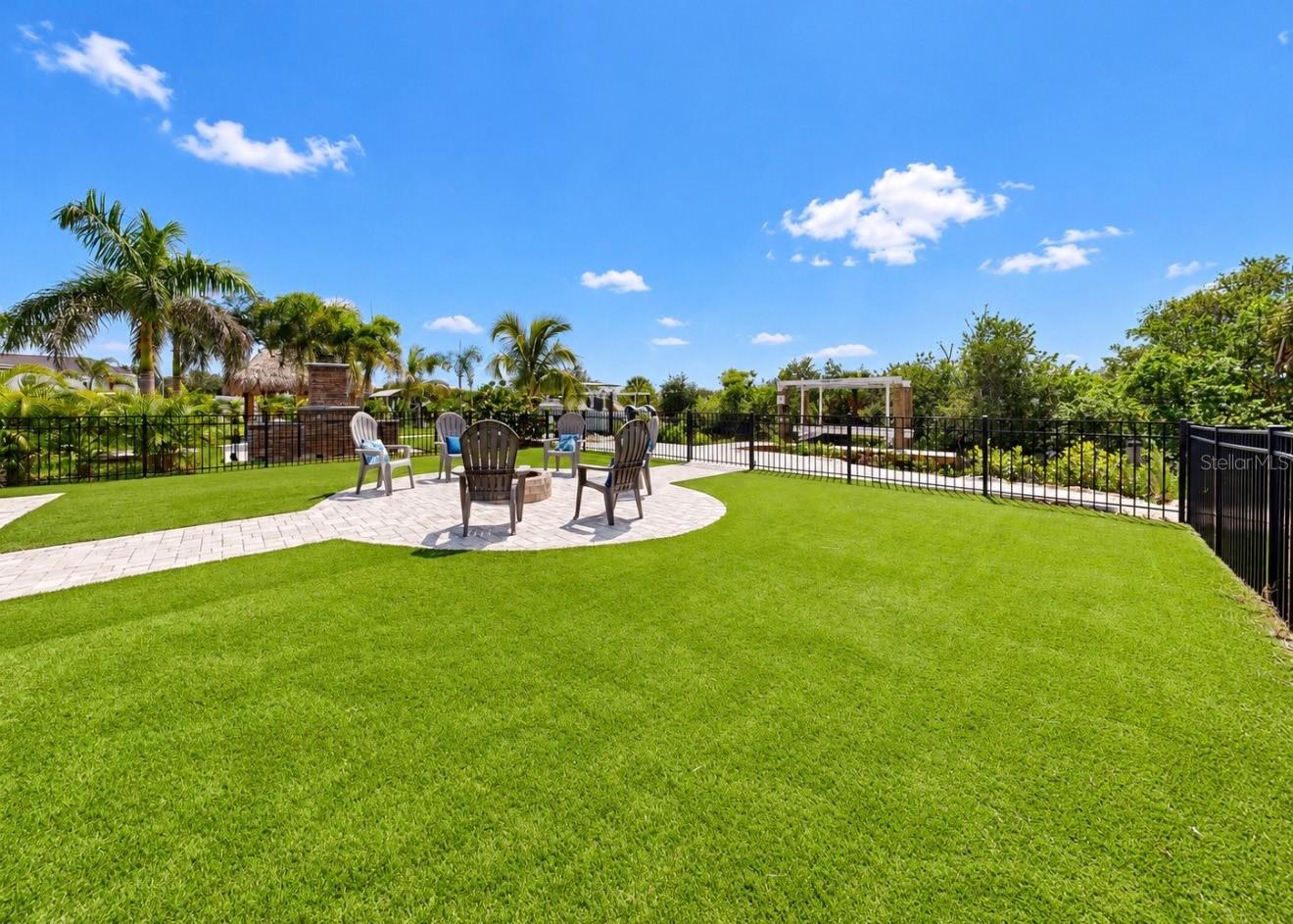 Landscaped backyard with a paver patio, Adirondack chairs, fire pit, palm trees and black metal fencing.
