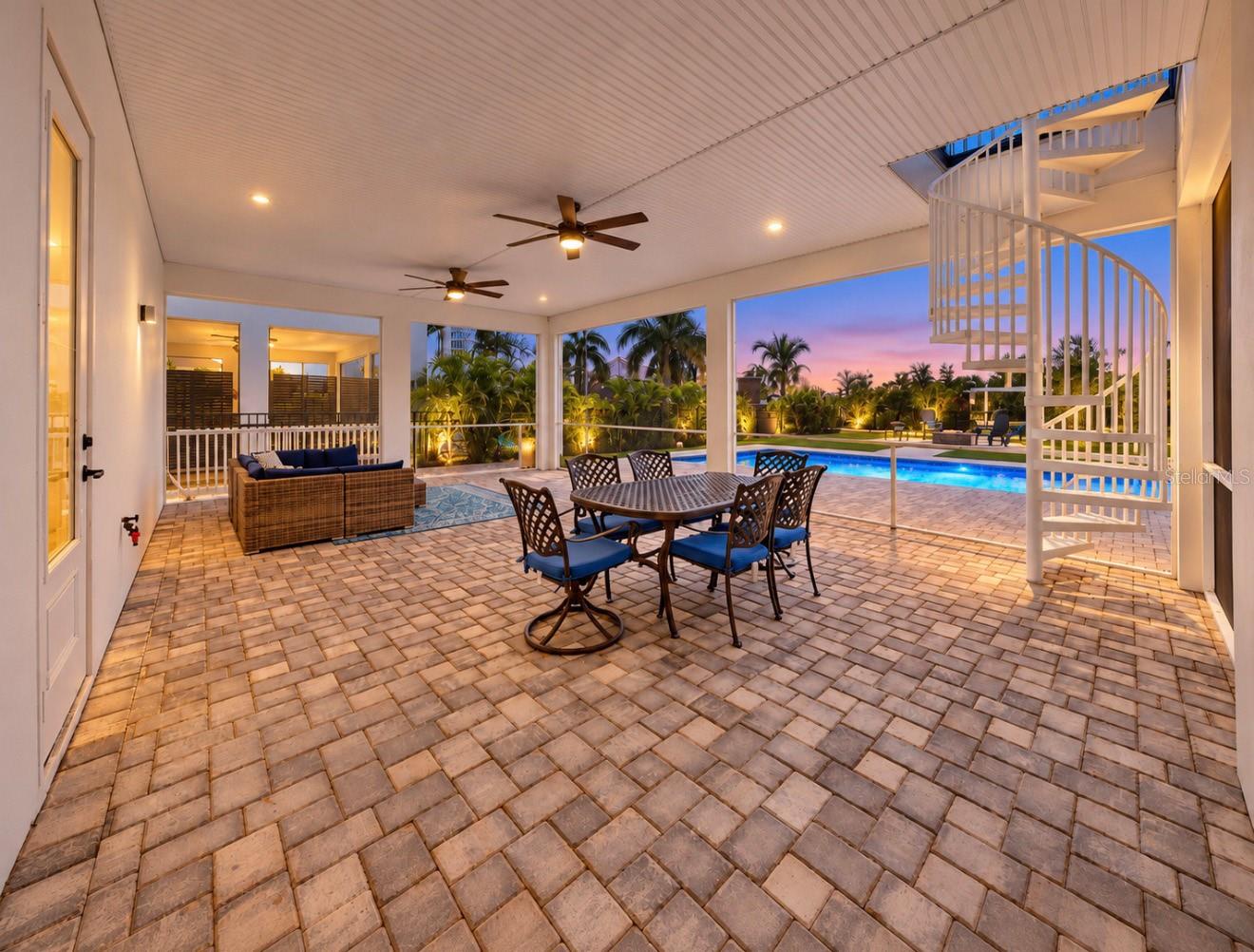 Covered paver patio with outdoor dining and lounge seating, a spiral staircase, and a pool at dusk.