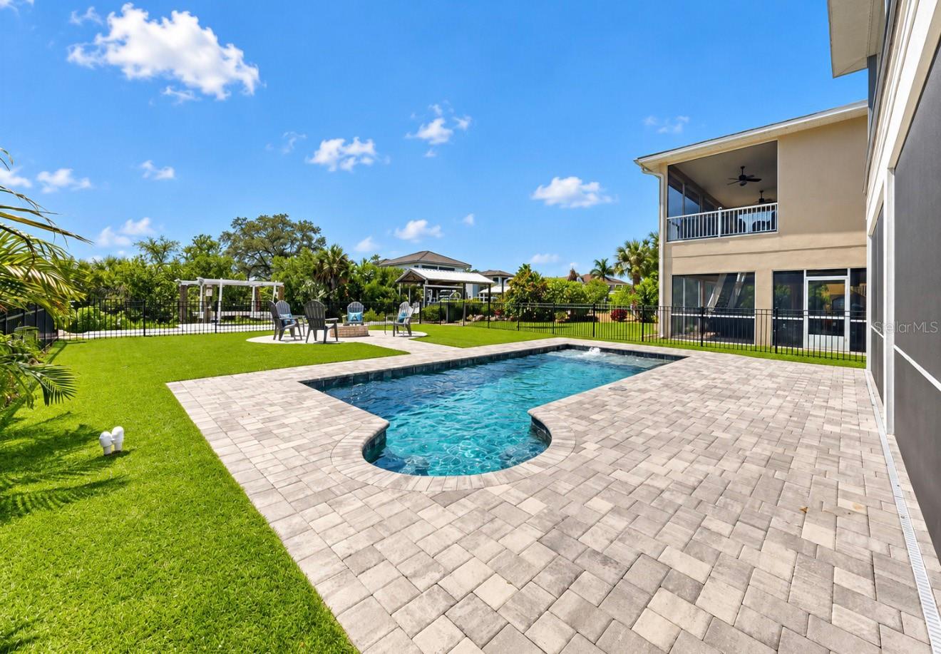 Backyard pool with a rounded end, broad paver patio, green lawn, and covered balcony.