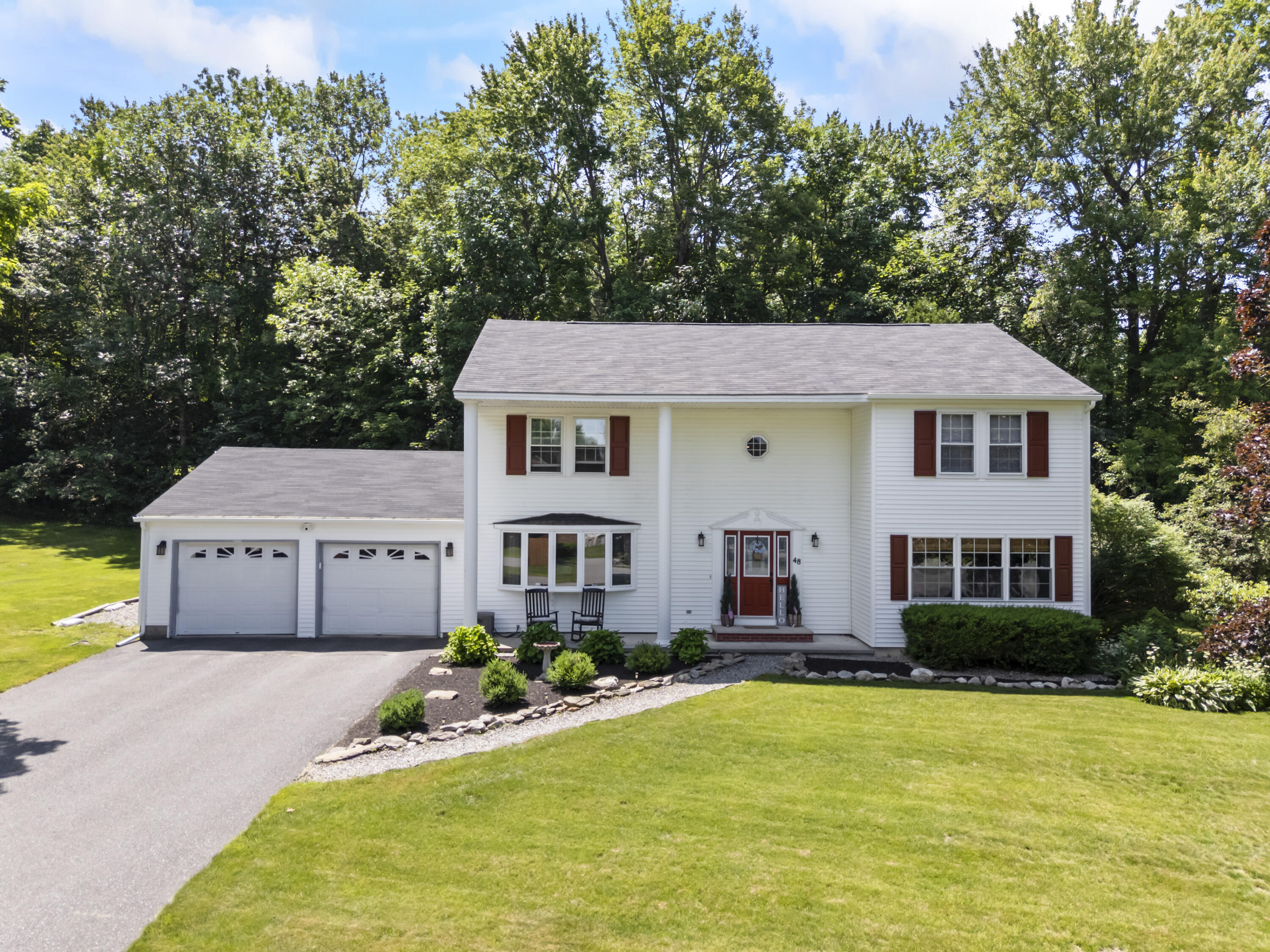 White two-story home with red shutters, attached two-car garage, and a wooded lawn.
