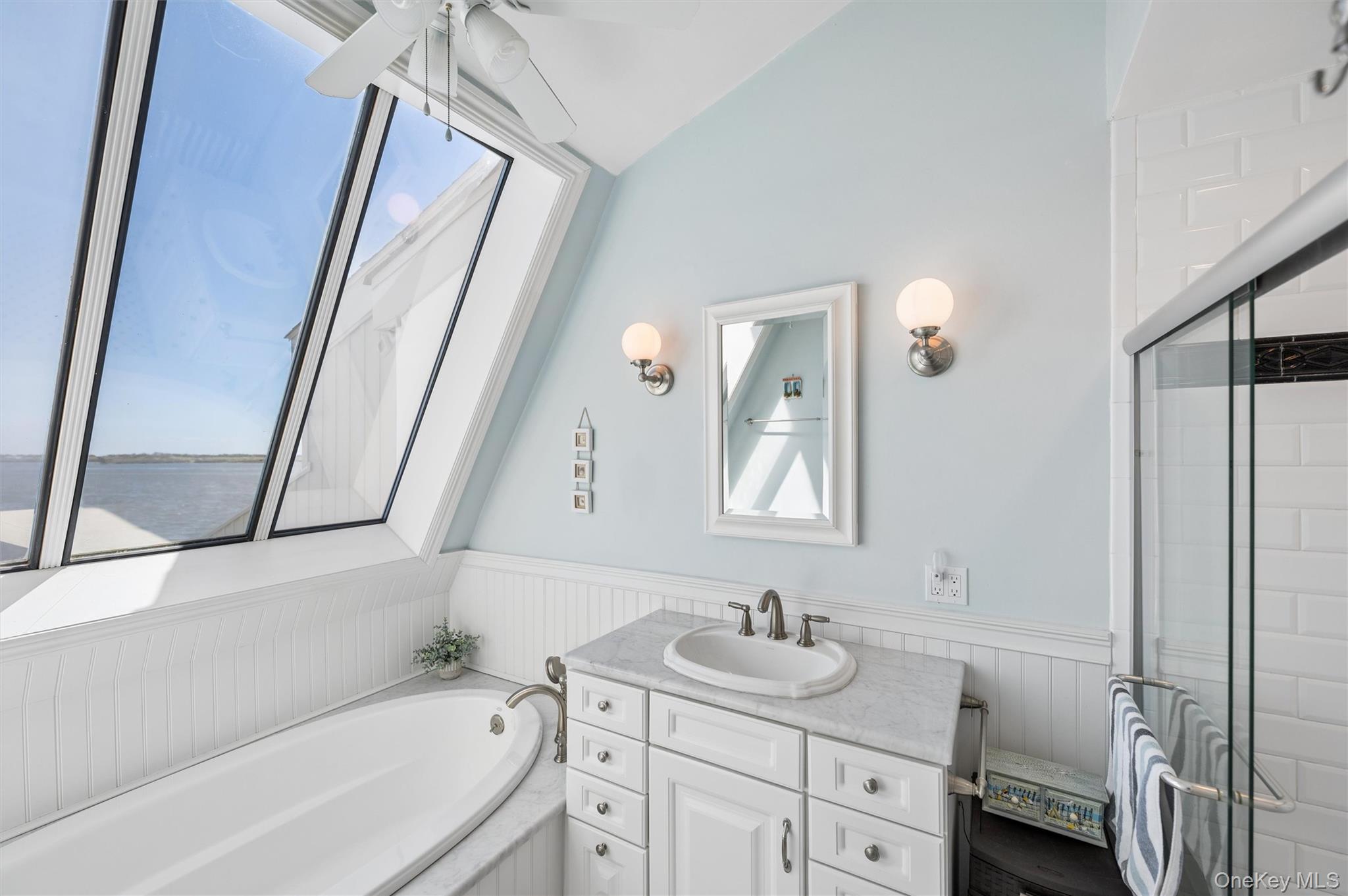 Bright bathroom with a soaking tub, marble vanity, glass shower, and slanted windows overlooking the water.
