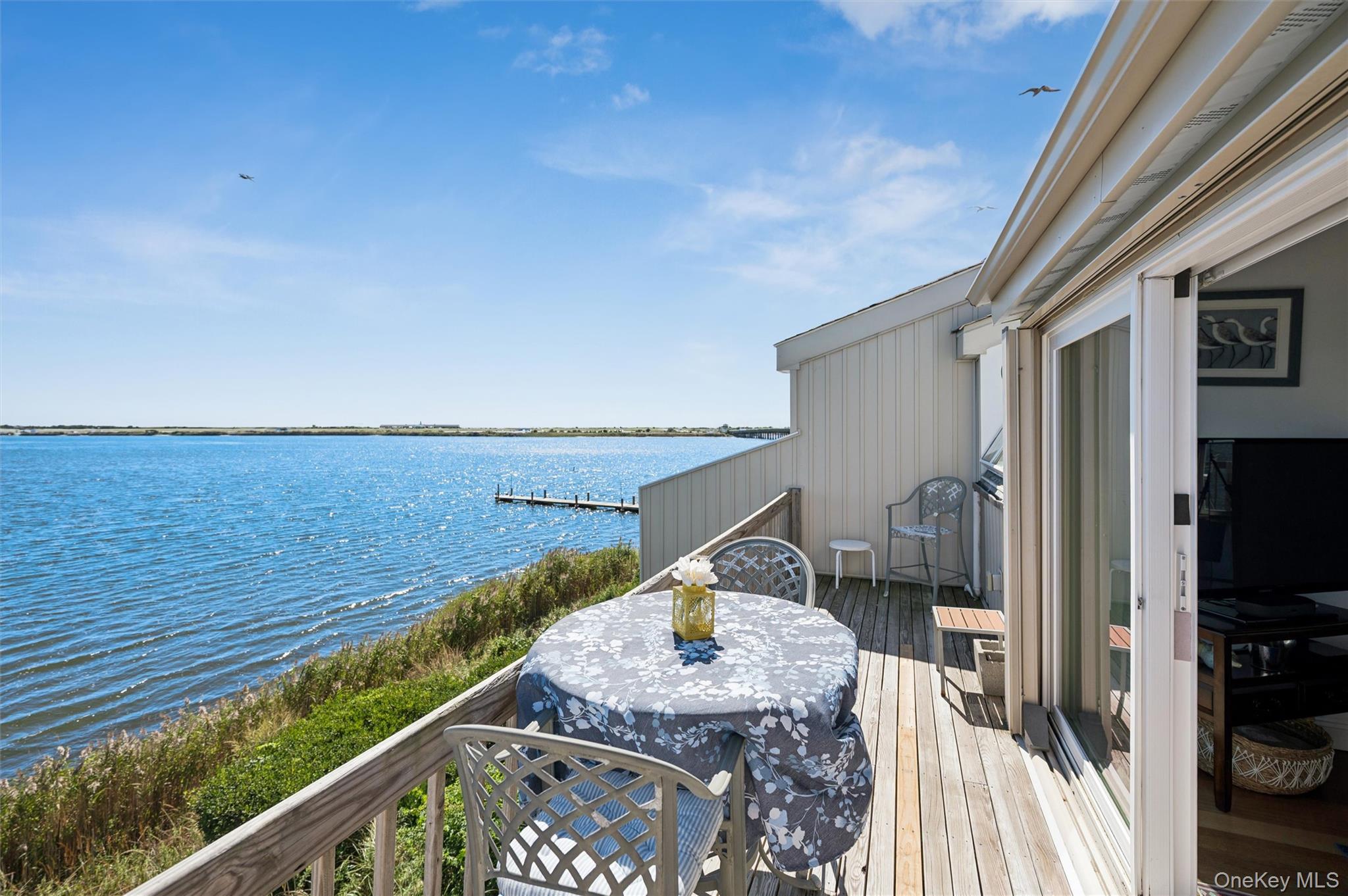 Waterfront deck with outdoor dining table overlooking blue water and a distant shoreline.