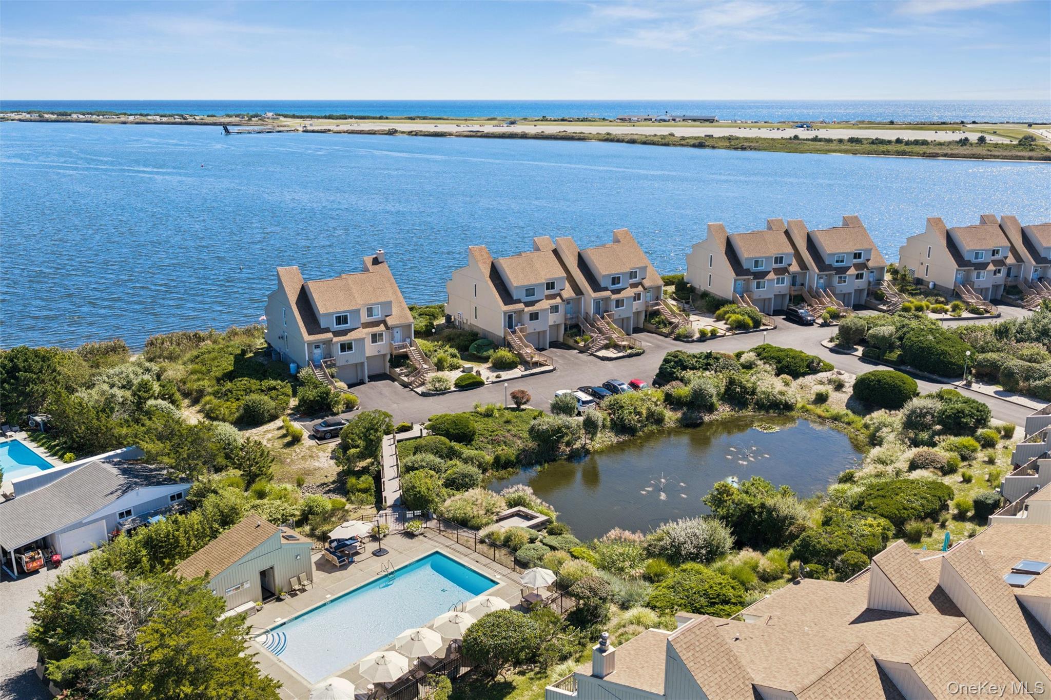 Aerial view of coastal townhouses beside a blue waterway, with a shared pool and landscaped pond.