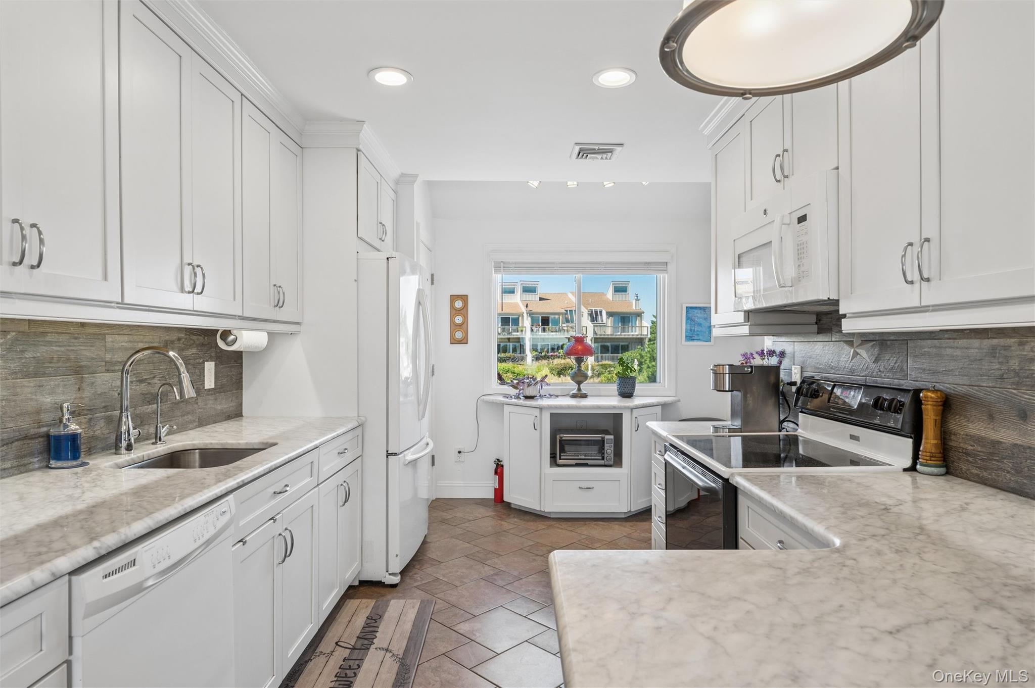Bright white kitchen with marble-pattern counters, stainless steel appliances, and a window overlooking neighboring homes.
