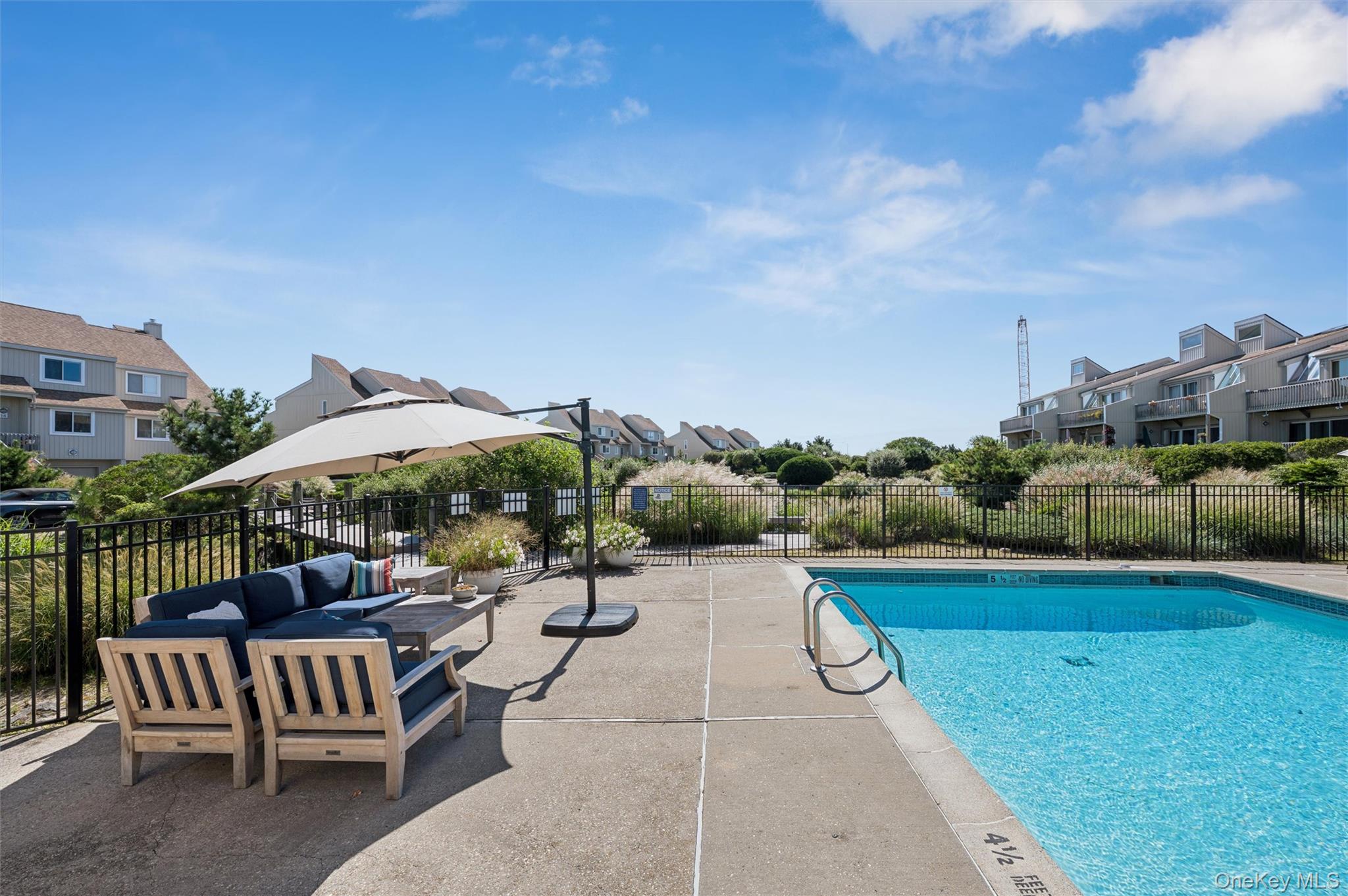 Outdoor swimming pool beside a furnished patio with umbrella-shaded seating and surrounding residential buildings.