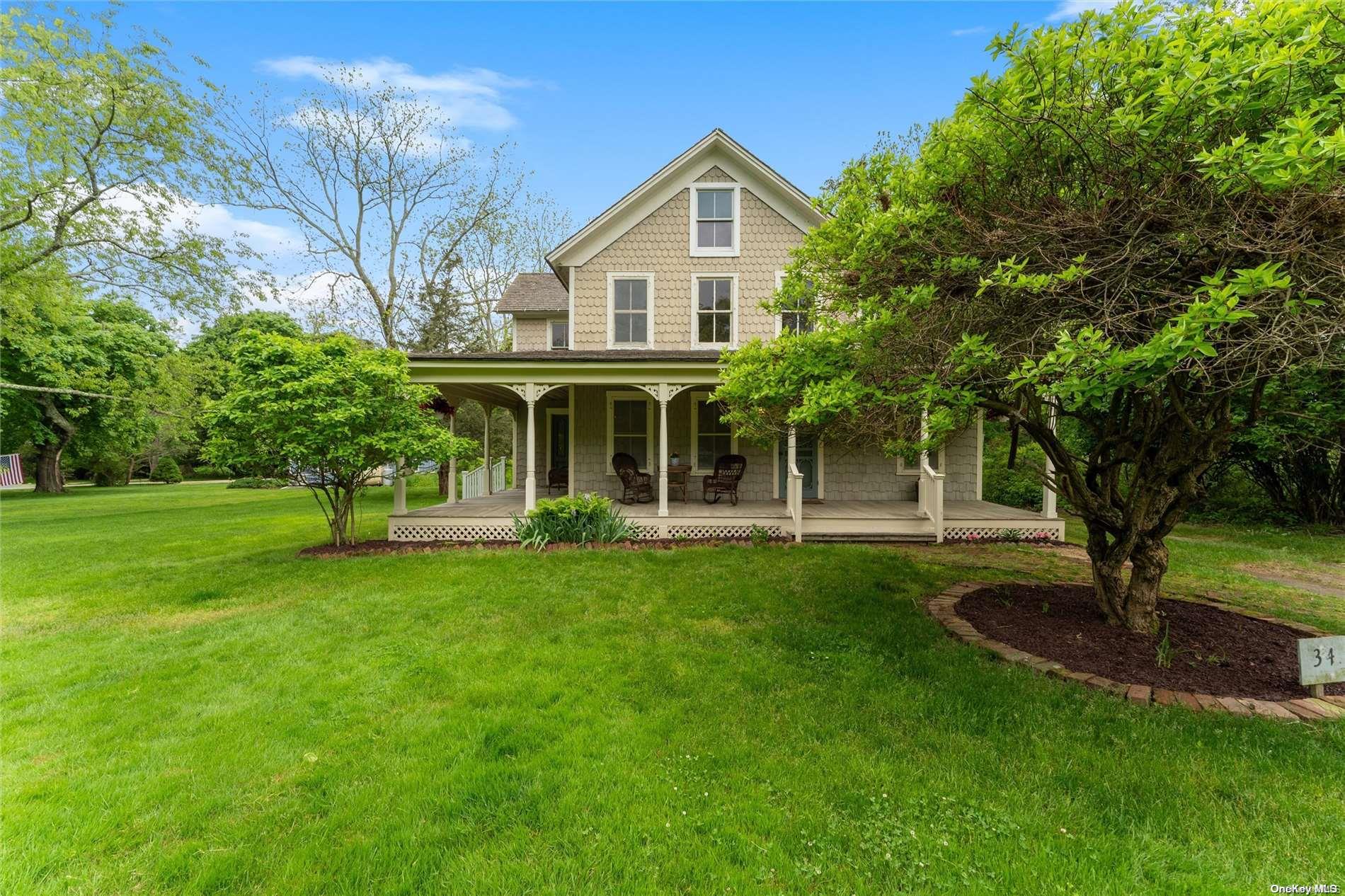 Shingle-sided two-story house with a covered porch and rocking chairs, set behind a broad green lawn.