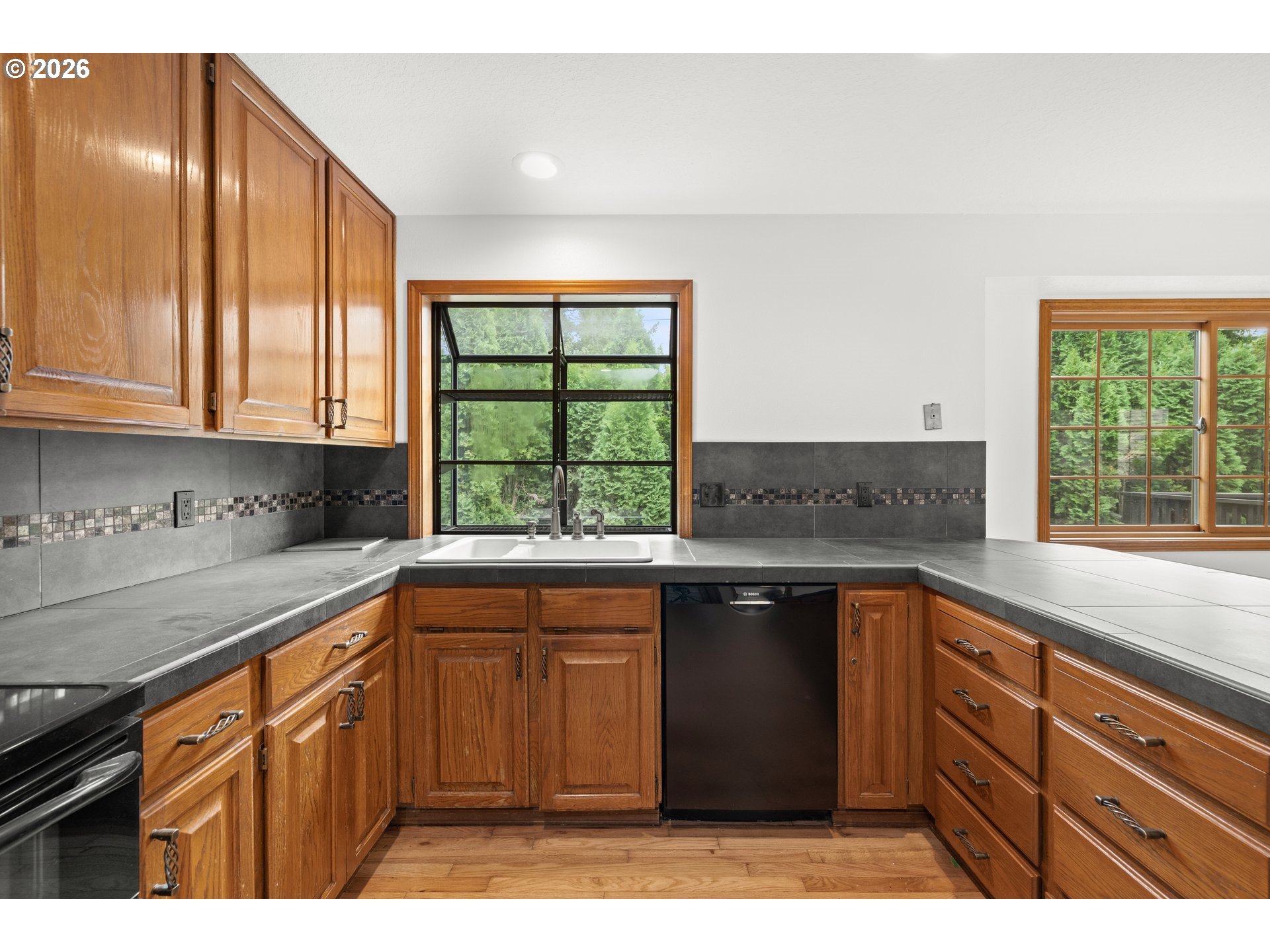 Kitchen with wood cabinets, gray tile counters, and a sink beneath a large window.