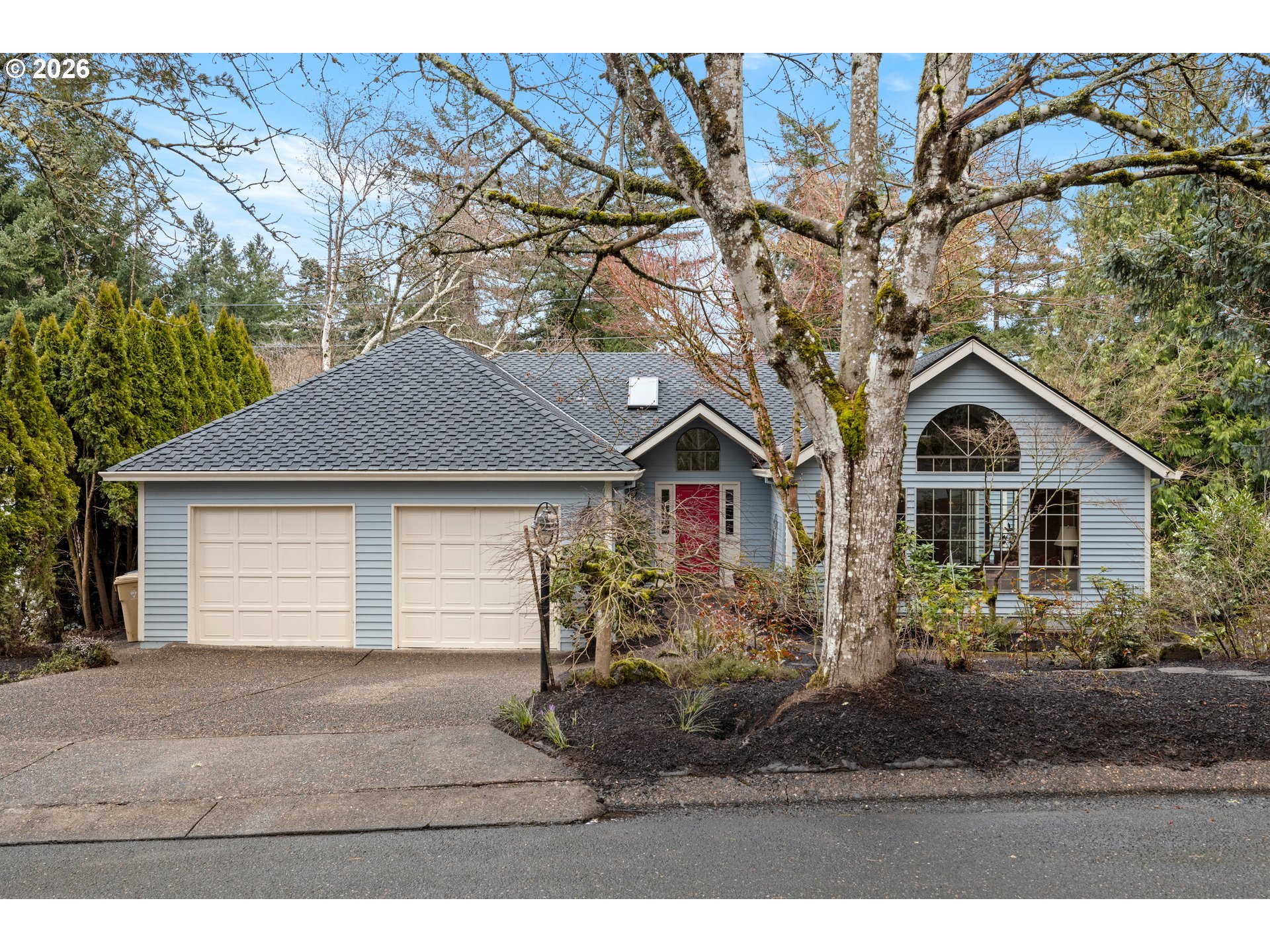 Blue-gray house with a red front door, two garage doors, and mature trees in the front yard.