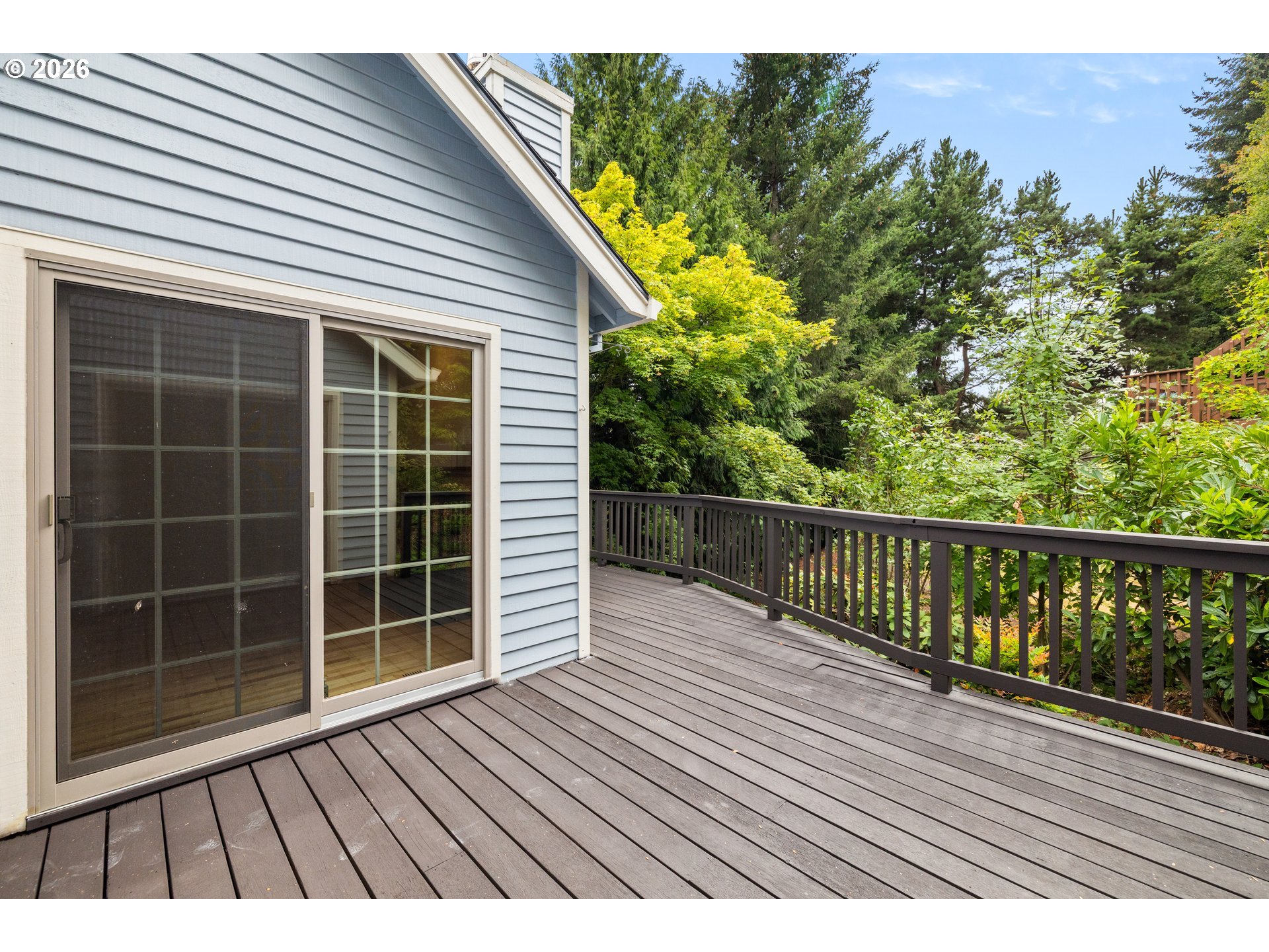 Spacious deck beside a blue-gray house, bordered by dark railings and dense greenery.