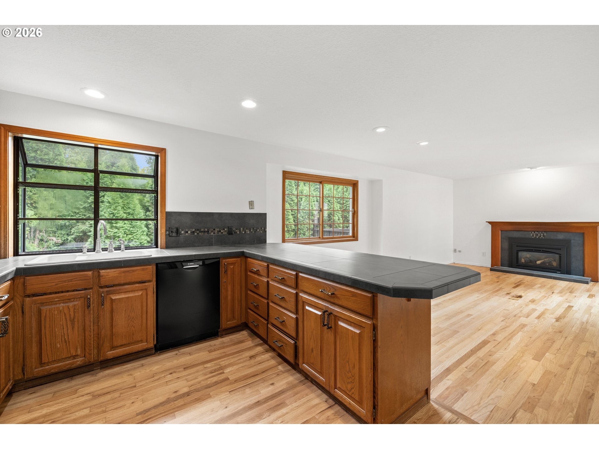 Kitchen with wood cabinets, dark tiled counters, large windows overlooking trees, and an adjoining fireplace.