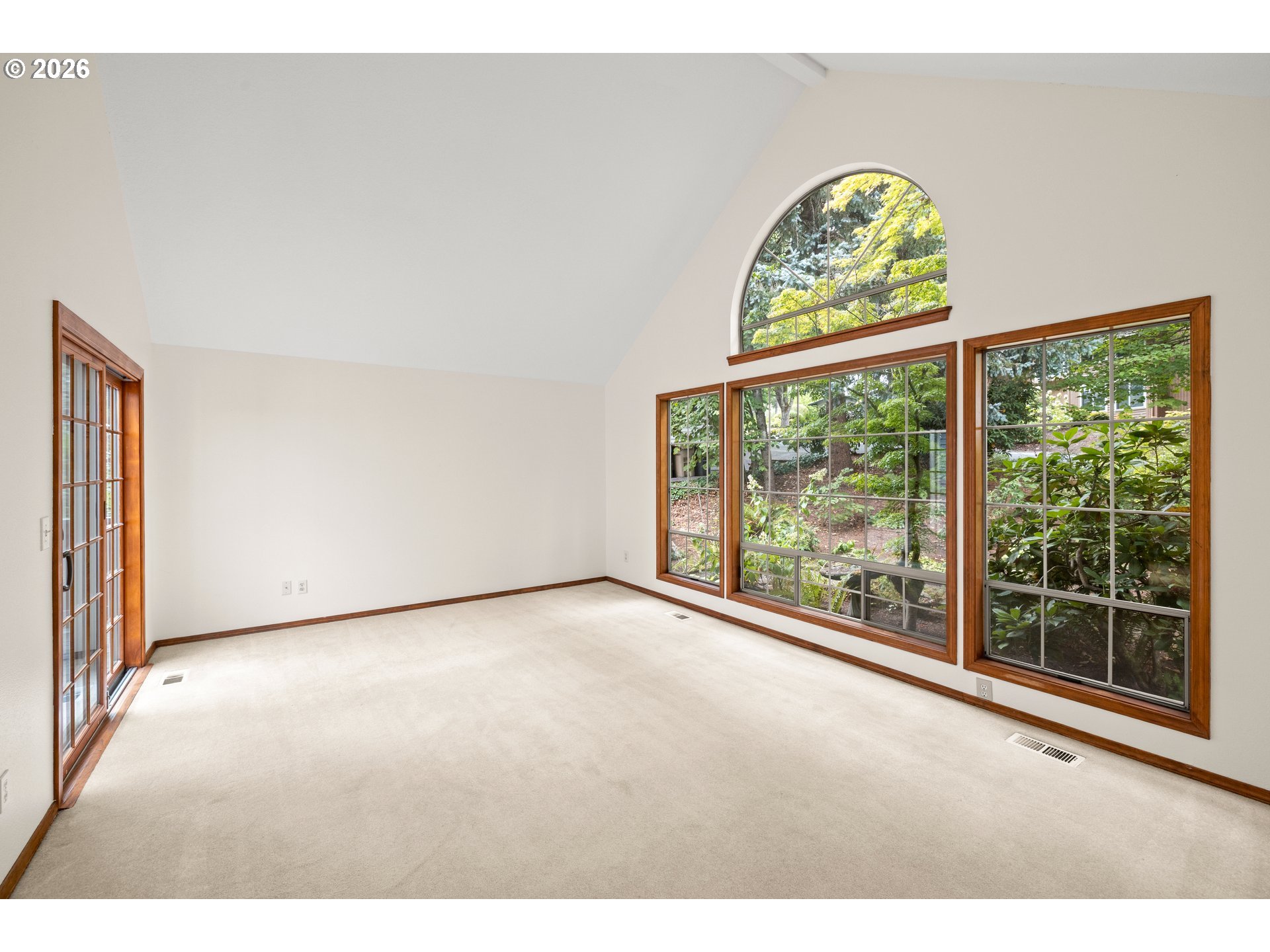 Empty carpeted room with a vaulted ceiling and large wood-trimmed windows overlooking greenery.