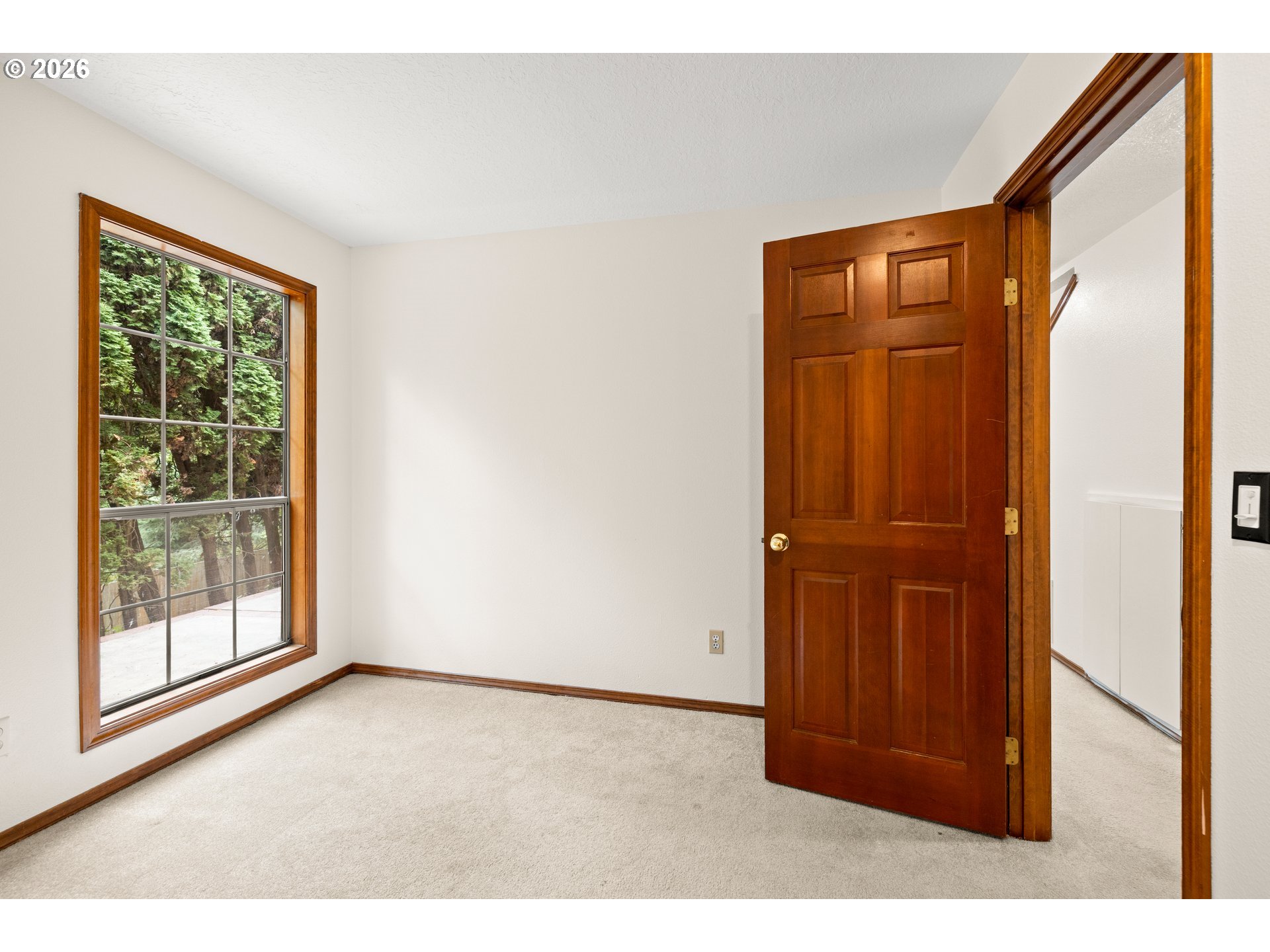 Empty carpeted room with a large window overlooking trees and an open wood-paneled door.