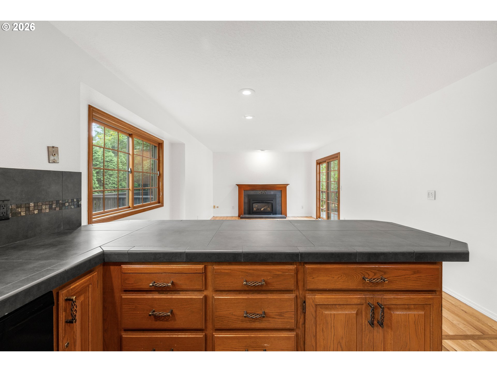 Kitchen island with dark tile countertop and wood cabinets, facing a living area with a fireplace.