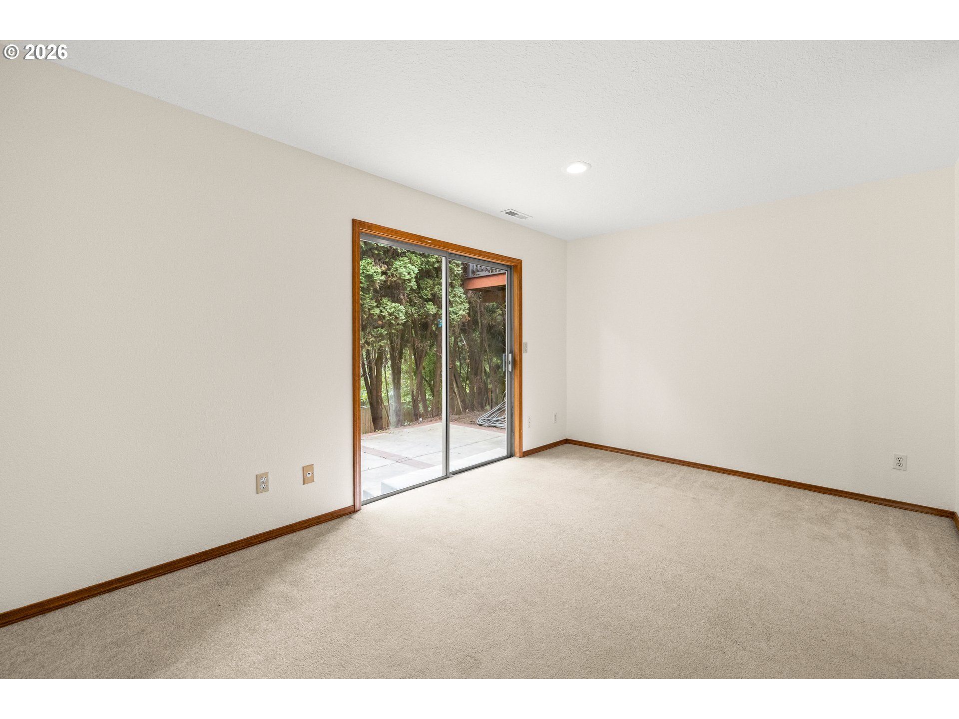 Empty carpeted room with a wood-trimmed sliding glass door overlooking trees.