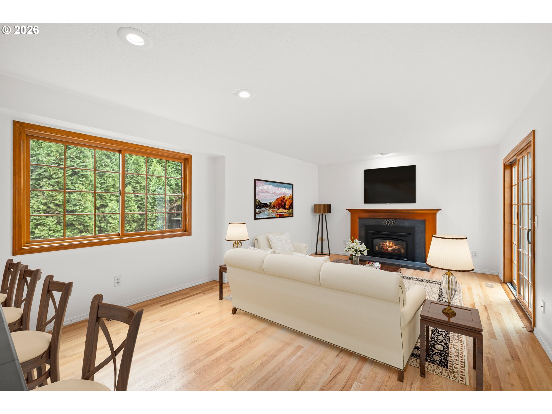 Living room with a wood-trimmed fireplace, white sofa, and large windows overlooking evergreen trees.