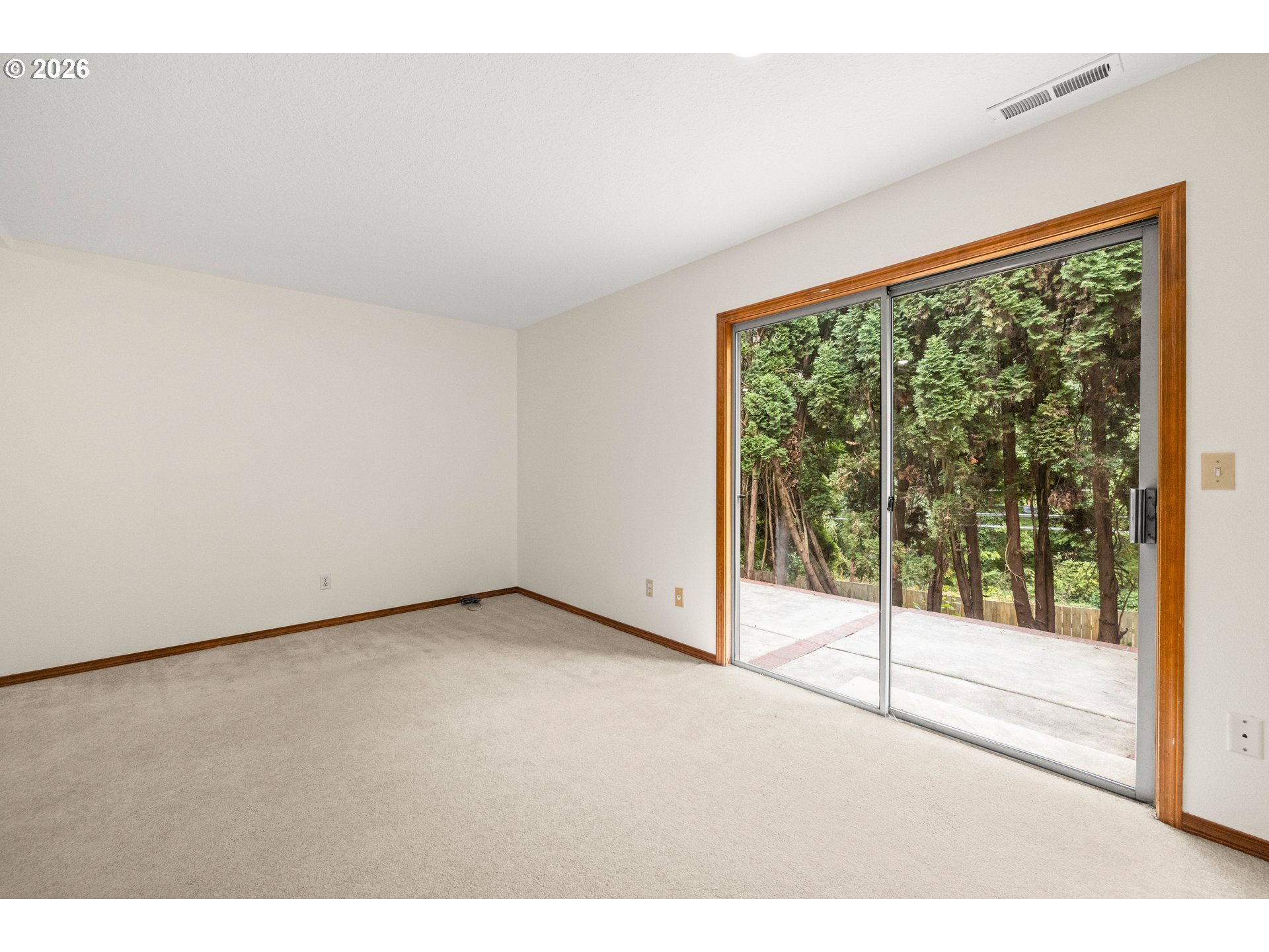 Empty carpeted room with wood-trimmed sliding glass doors overlooking evergreen trees.
