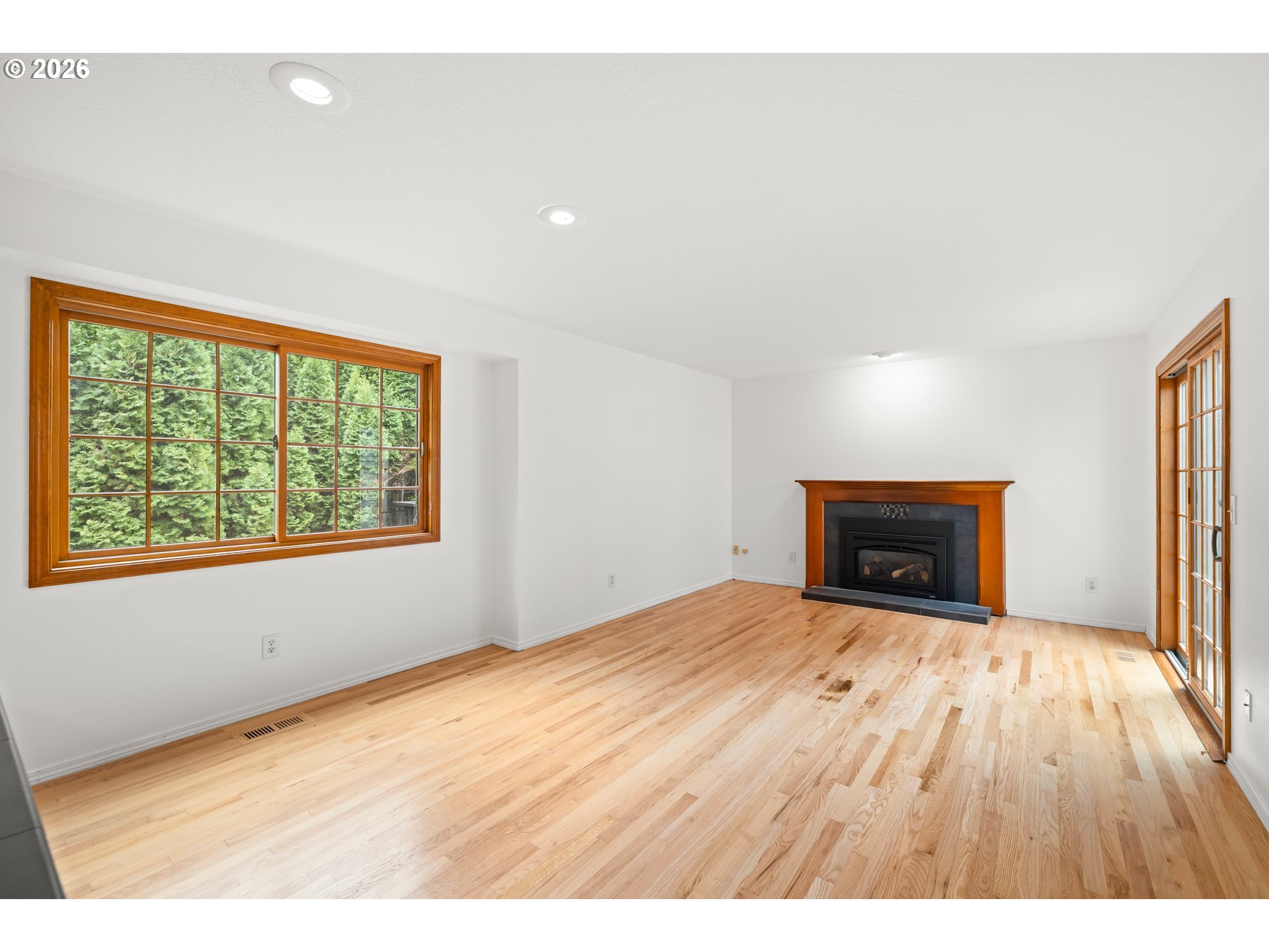 Vacant living room with light hardwood floors, a wood-trimmed fireplace, and large windows overlooking evergreens.
