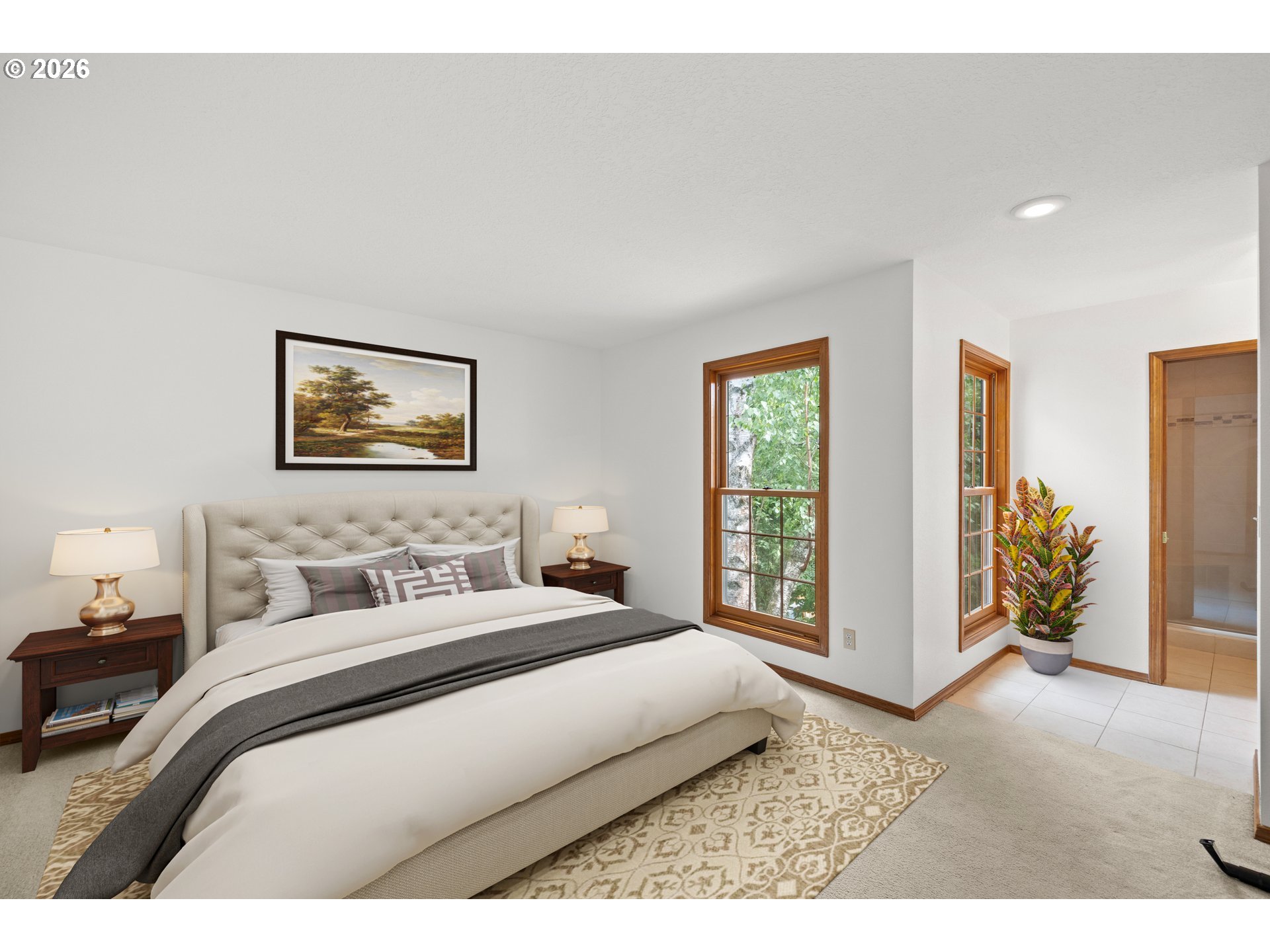 Staged bedroom with a cream bed, patterned rug, and wood-trimmed windows overlooking leafy trees.