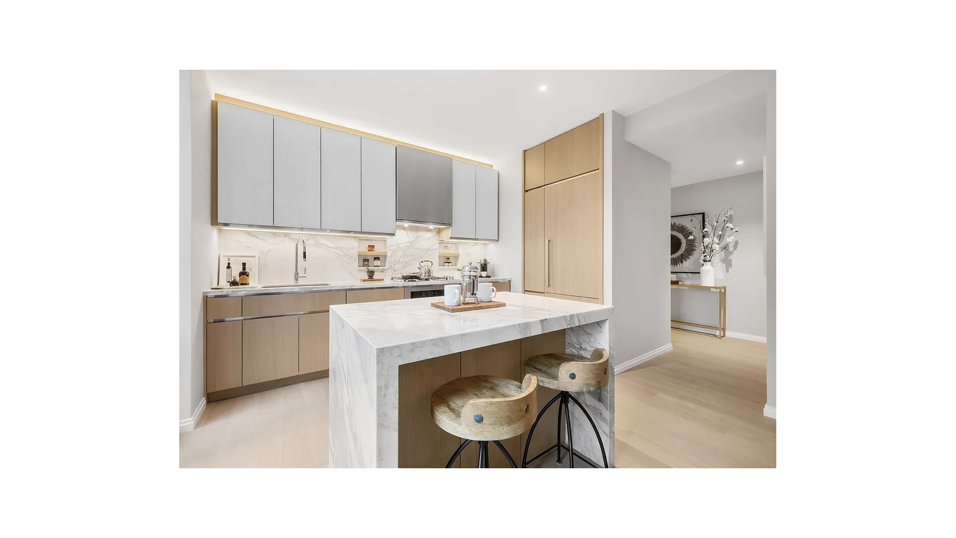 Modern kitchen with a large marble island, two stools, and pale gray and wood cabinetry.
