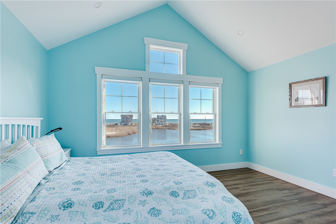 Bedroom with vaulted ceiling, coastal-pattern bedding, and three windows overlooking water.
