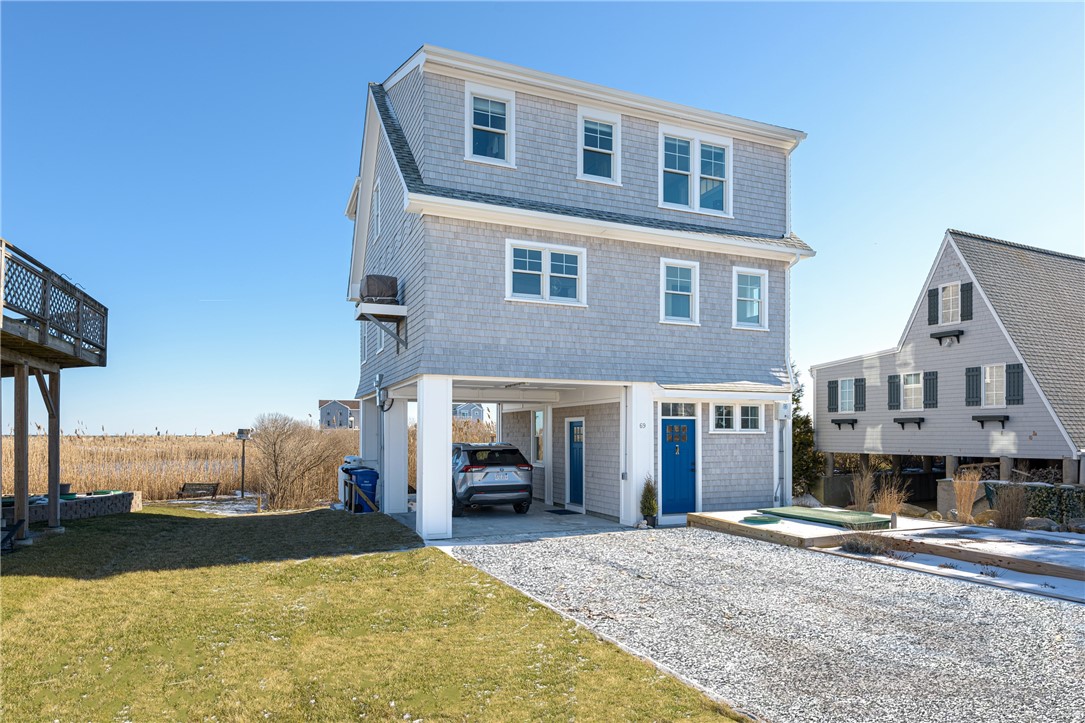 Gray shingle-sided coastal home with a blue door, covered carport, and gravel driveway.