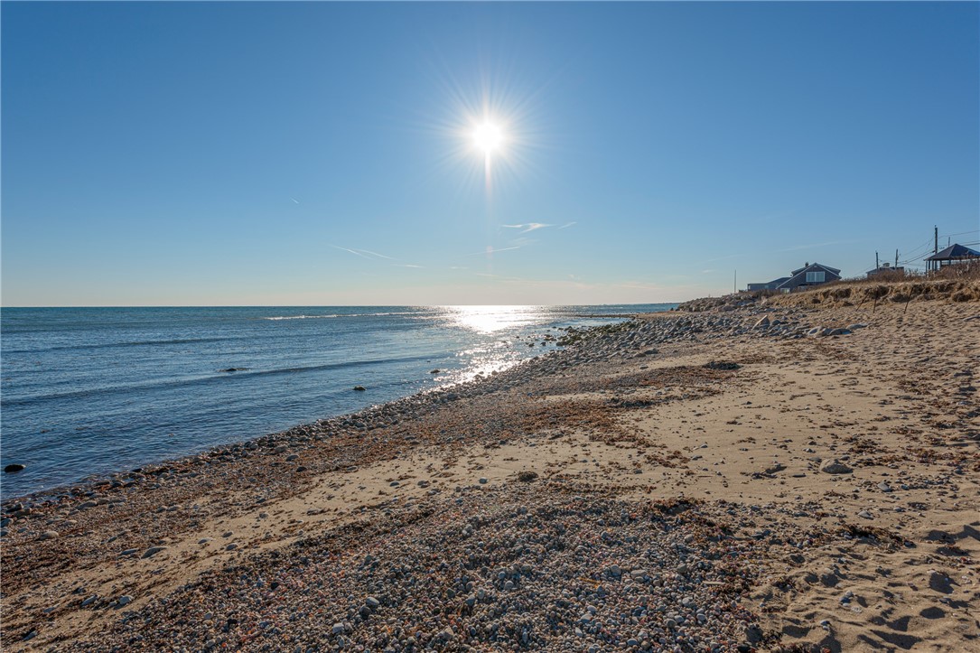 Rocky beach and ocean shoreline beside distant homes under bright sunlight.
