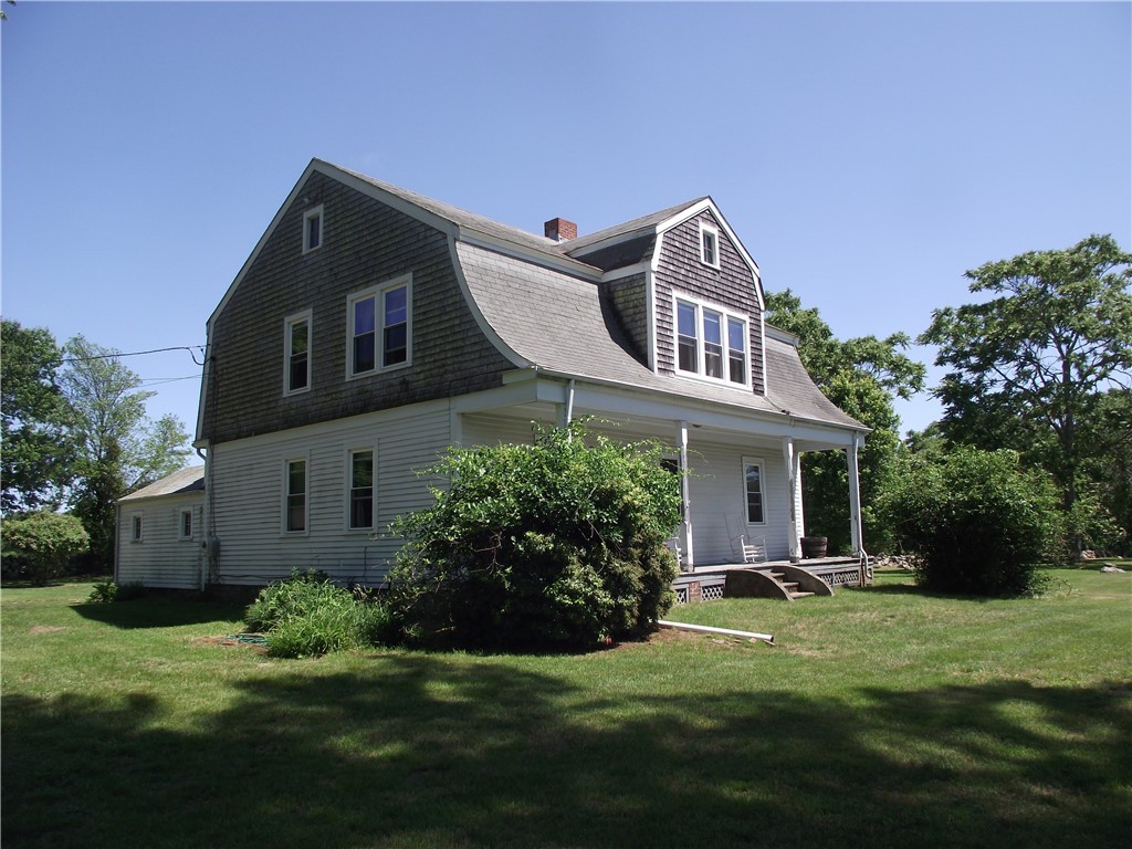 Gray-shingled gambrel-roof house with a covered porch on a sunny lawn.
