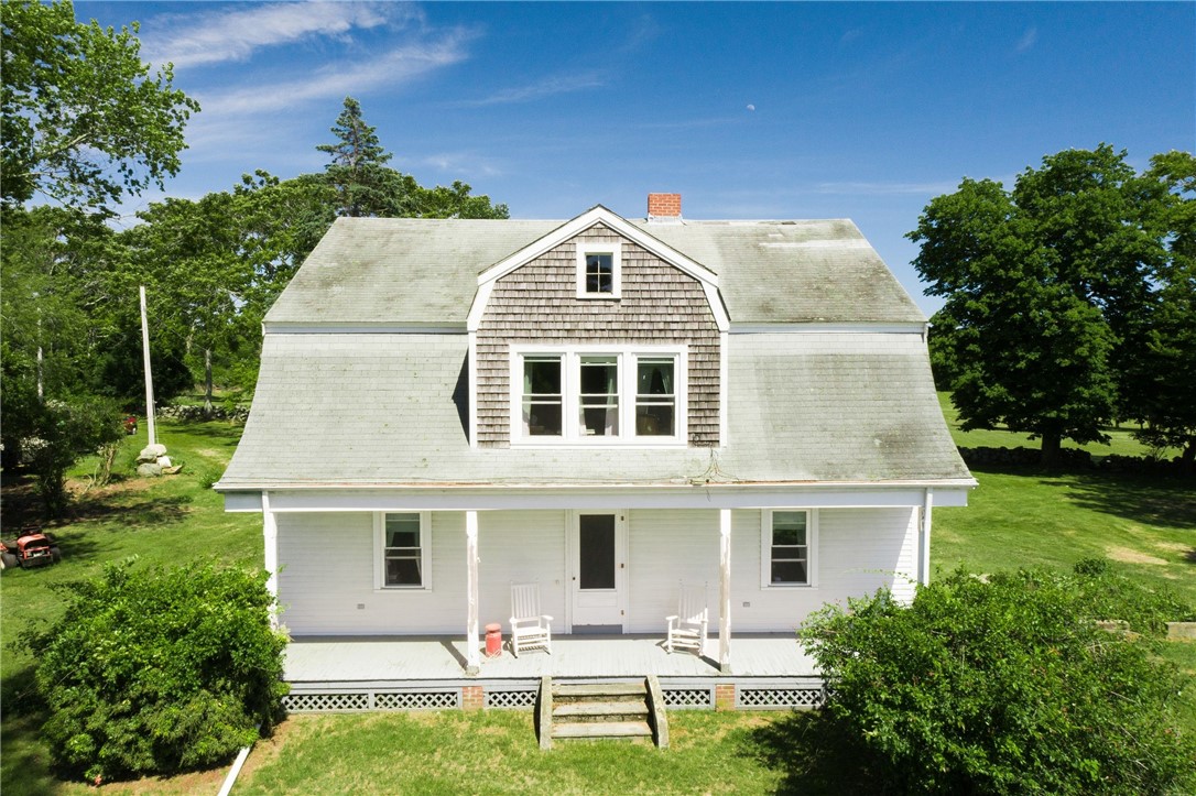 White two-story house with a gambrel roof, front porch, and two chairs on a sunny tree-lined lawn.