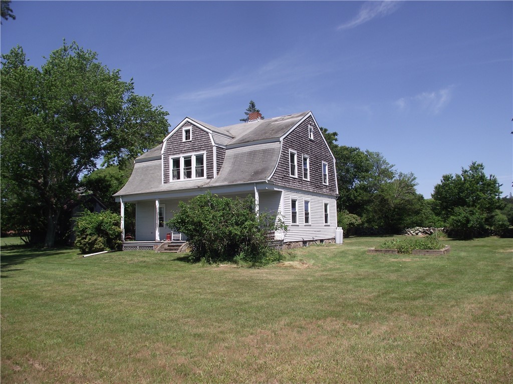 Gray-shingled two-story house with a gambrel roof and covered porch in a broad grassy yard.