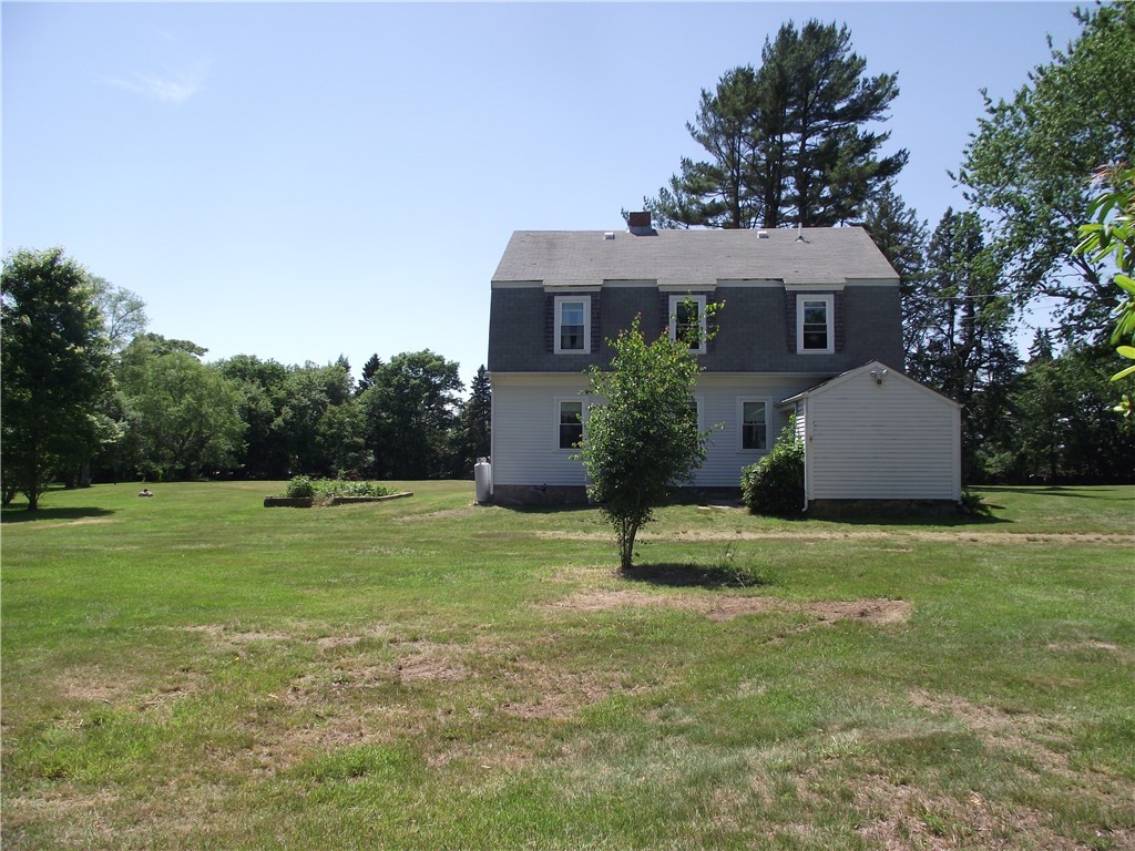Rear of a two-story gray house with a side addition, surrounded by a broad lawn and mature trees.