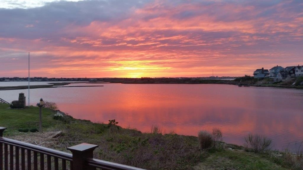 Coastal inlet glowing pink beneath a sunset, with shoreline homes and a grassy bank.