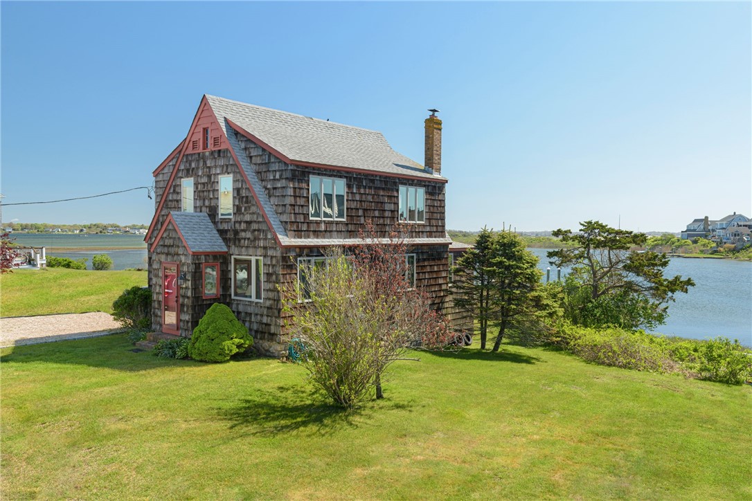 Cedar-shingle cottage with red trim on a waterfront lawn beside an inlet.