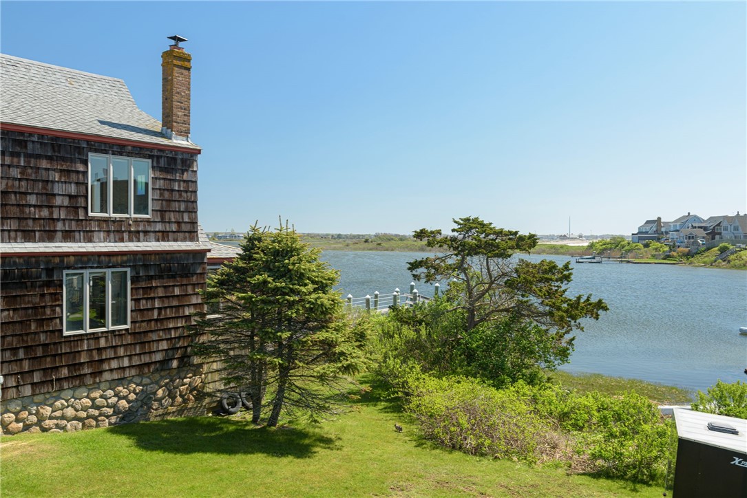 Shingle-sided coastal home beside an inlet, with a stone foundation and lawn leading to the water.