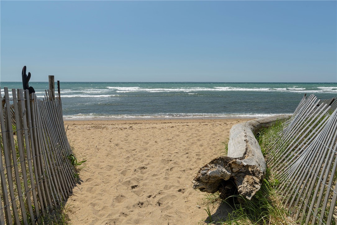 Sandy beach access between weathered dune fences, opening onto the ocean beside a large piece of driftwood.