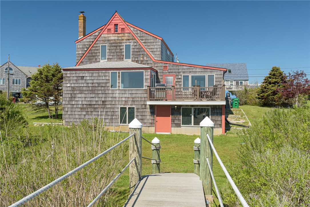Coastal shingle house with coral-red trim, raised deck, and wooden walkway leading to the entrance.