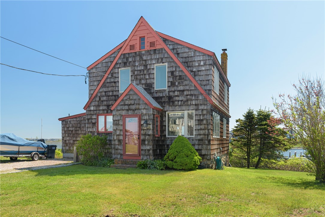 Shingle-sided home with red trim and steep gables, a green lawn, and water nearby.