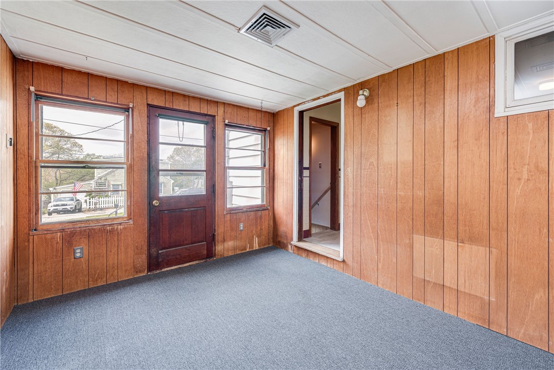 Empty wood-paneled room with a carpeted floor, windowed exterior door, and open doorway to a stairwell.