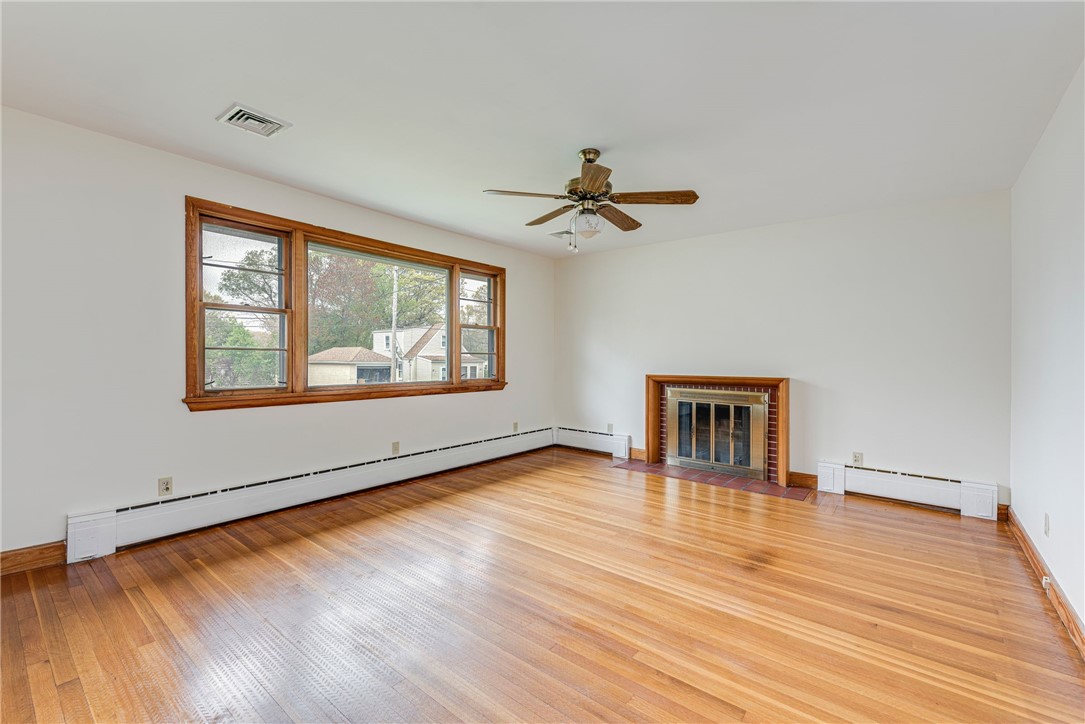 Empty living room with hardwood floors, a wide window, brick fireplace, and ceiling fan.