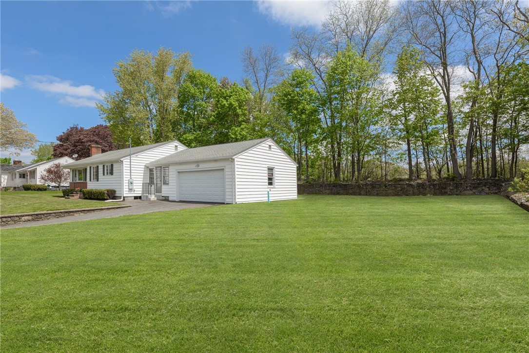White single-story house with attached garage beside a broad lawn and tree-lined stone wall.