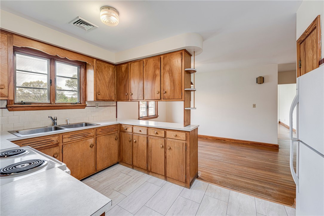 Kitchen with wood cabinetry, a double sink under two windows, and an opening to a hardwood-floored room.