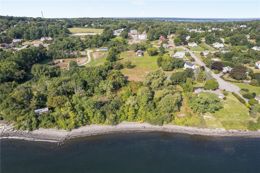 Aerial view of a wooded neighborhood along a rocky shoreline, with homes and open land near the water.