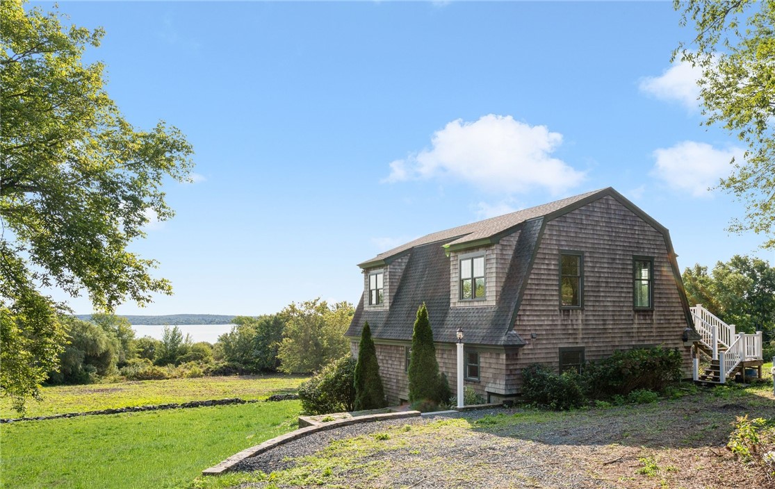 Cedar-shingle home with a gambrel roof, set above a lawn with water visible beyond the trees.